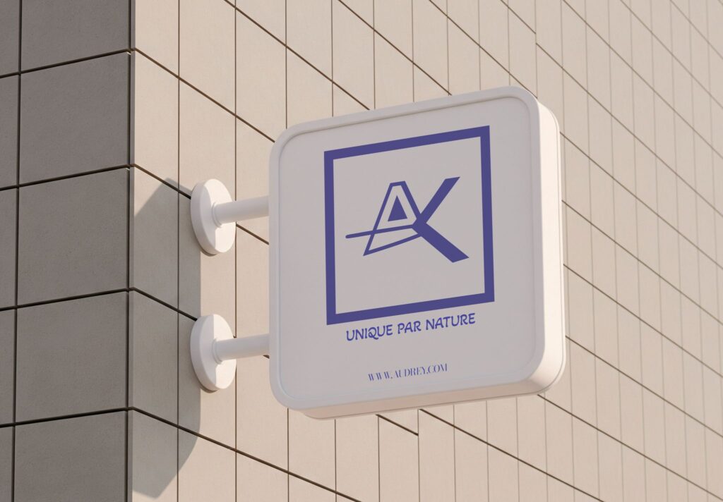 Mockup enseigne murale du logo AK - monogramme bleu indigo sur fond blanc, version carré encadré, fixé sur un mur de façade beige carrelé. Tagline "Unique par Nature" et URL www.audrey.com en dessous. Rendu 3D réaliste et élégant qui met en valeur l'identité visuelle personnelle d'Audrey.