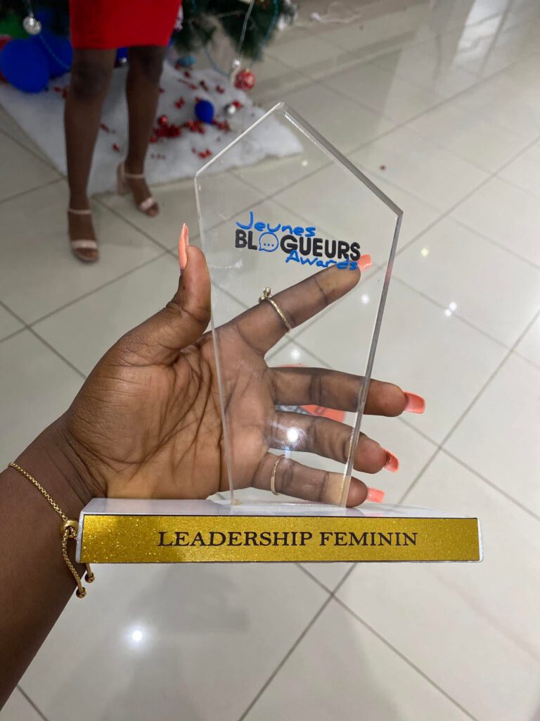 Photo du trophée "Jeunes Blogueurs Awards - Leadership Féminin" tenu dans la main d'Audrey aux ongles corail et bracelet doré. Trophée en plexiglas transparent en forme de maison stylisée, sur socle blanc et doré pailleté. Une belle récompense méritée !