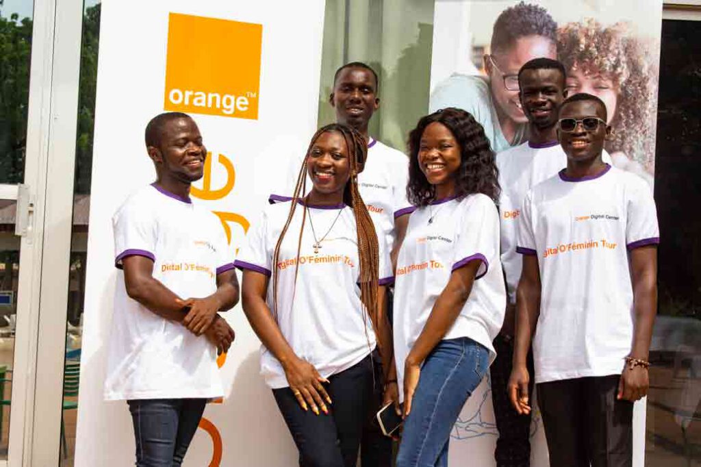 Photo de groupe du Digital O'Féminin Tour en partenariat avec Orange Digital Center - cinq jeunes membres de l'équipe souriants, vêtus de t-shirts blancs à liseré violet floqués "Digital O'Féminin Tour", posent devant un kakémono Orange. Deux femmes aux longs tresses au centre, entourées de trois hommes. Ambiance joyeuse et soudée, témoignant de l'esprit d'équipe derrière cet événement de formation numérique au féminin en Côte d'Ivoire.