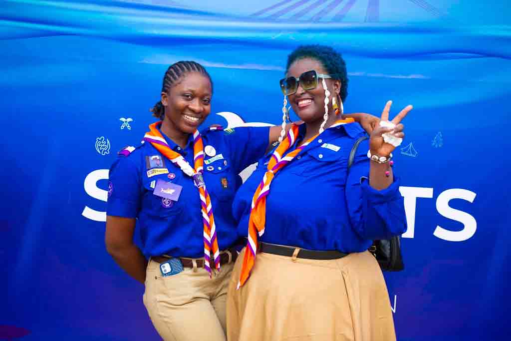 Photo de deux scouts souriantes en uniforme bleu royal avec foulard orange et blanc, posant devant un fond bleu lors d'un événement scout. L'une porte plusieurs badges et insignes sur sa chemise, l'autre arbore des lunettes de soleil tendance et fait le signe de la victoire. Ambiance joyeuse et conviviale.