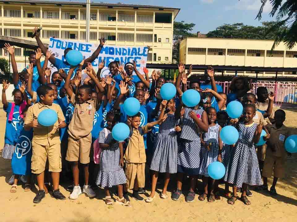 Photo de la Journée Mondiale de l'Enfance organisée par les Jeunes Blogueurs - UNICEF Côte d'Ivoire - grand groupe d'enfants en uniforme scolaire et bénévoles en t-shirts bleus UNICEF, les mains levées en signe de joie, tenant des ballons bleus. Ambiance festive et colorée dans la cour d'une école, avec une banderole "Journée Mondiale de l'Enfance" accrochée en arrière-plan. Un moment fort célébrant les droits et le bonheur des enfants.